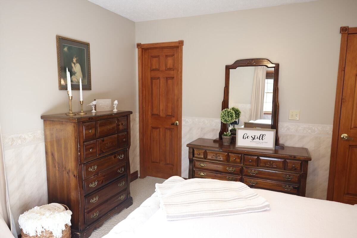 A bedroom with a wooden dresser, a bed with white linens, a mirror, a door, and a framed picture on the wall.