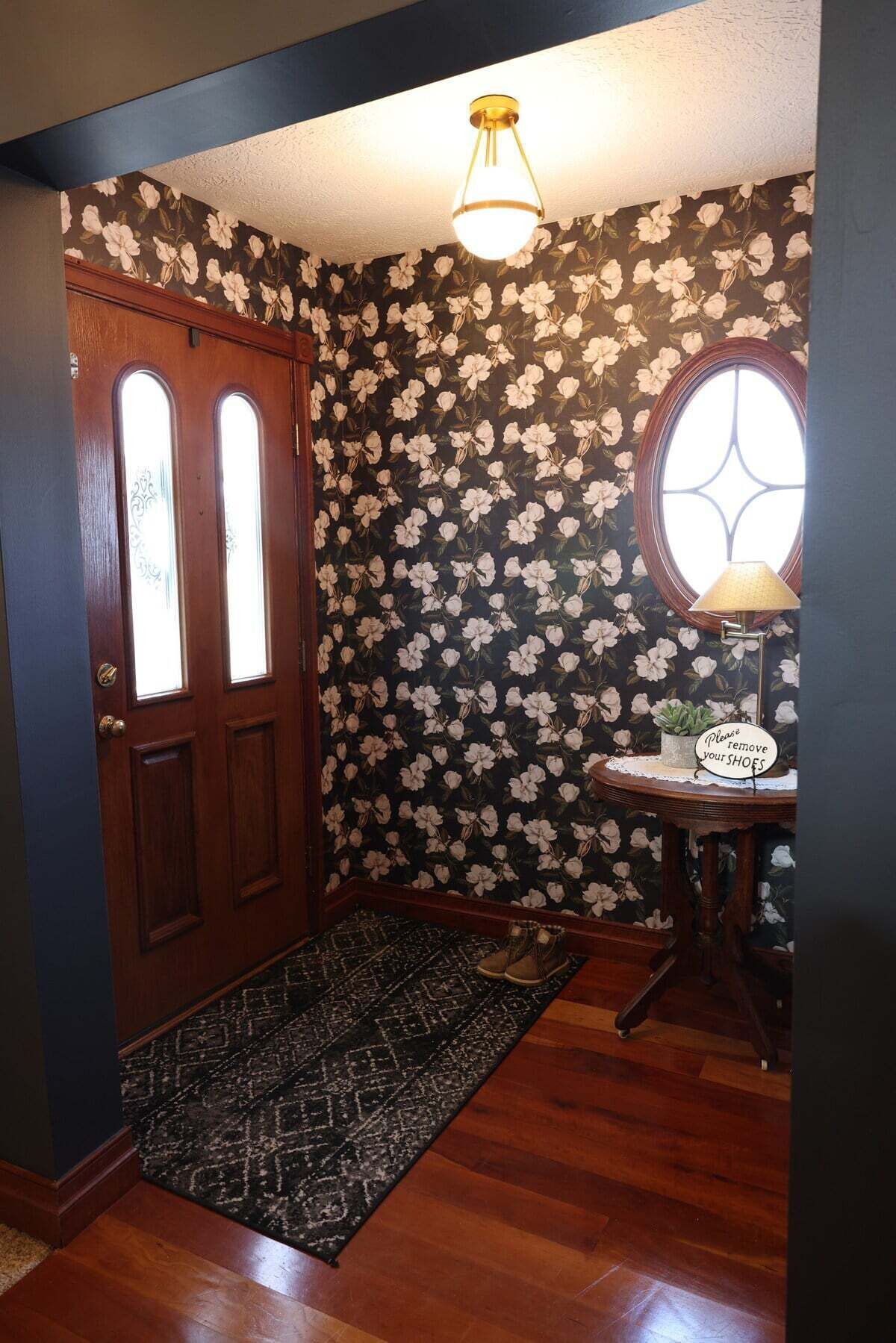 Una porta in legno con un pannello di vetro, una parete scura con motivo decorativo, una finestra tonda, un tavolino con una lampada e un tappeto.