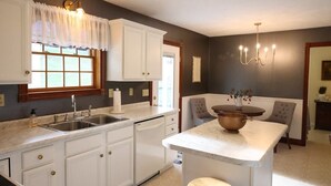 A kitchen with white cabinets, a sink, a dishwasher, a marble countertop, a chandelier, and a dining area with chairs. - Spacious Family Home w/ Fireplace & Fire Pit (Grayson)