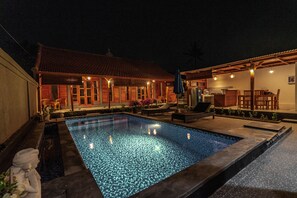 A swimming pool with a tiled deck, a covered seating area with chairs and tables, and a building with a tiled roof and windows.