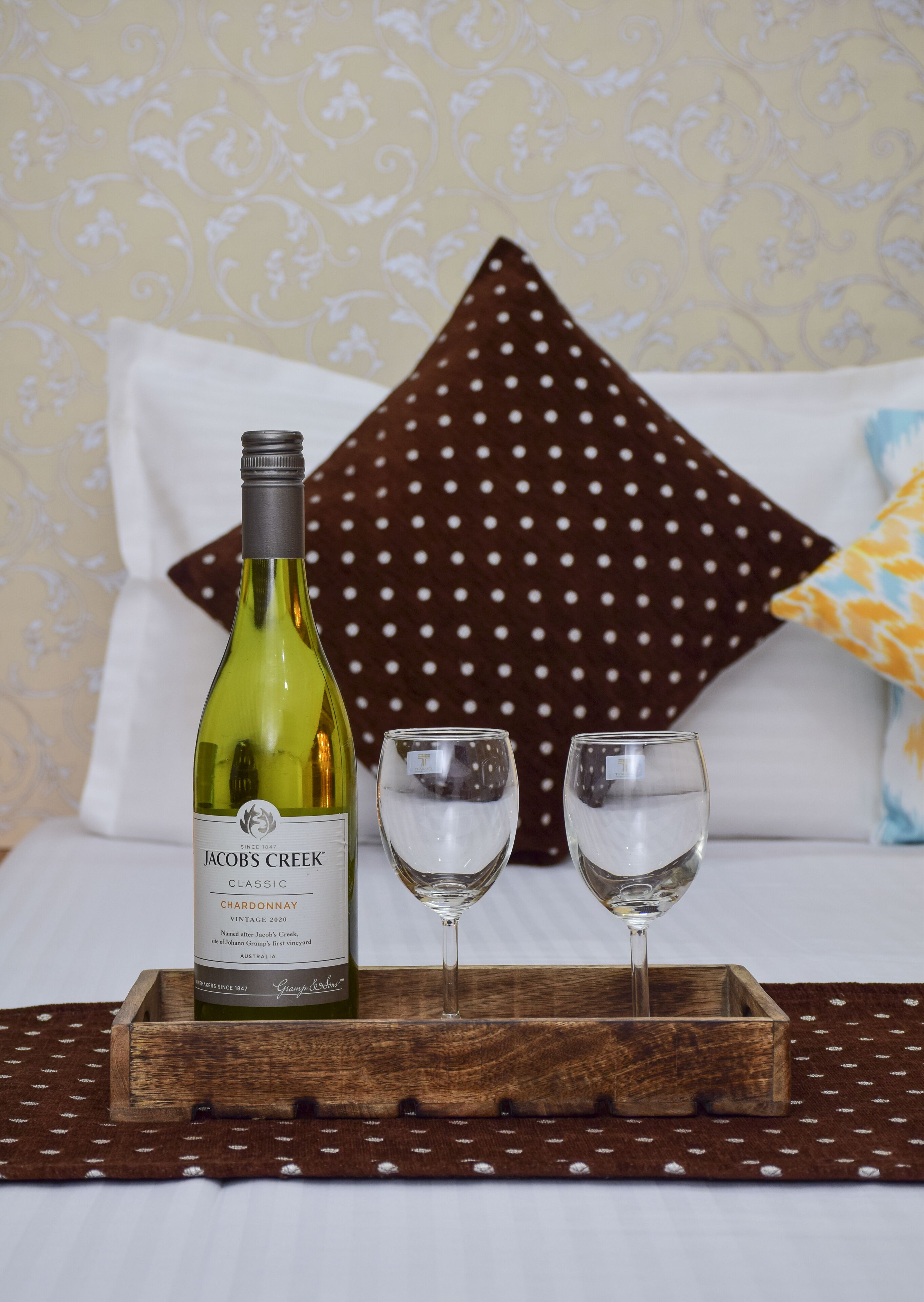 Una botella de Jacob's Creek Chardonnay sobre una bandeja de madera con dos copas, colocada sobre una cama con almohadones estampados.