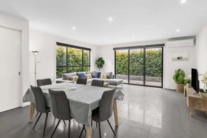 Uma sala de jantar moderna com mesa e cadeiras, um sofá com almofadas, e grandes janelas com vista para o verde.