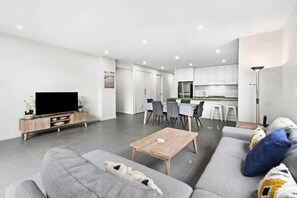 Una sala de estar moderna con un sofá, una mesa de centro y un televisor. Se observa un comedor con sillas y una cocina al fondo.