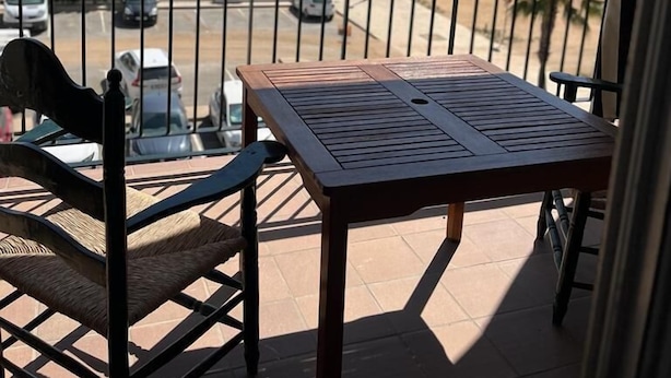 A balcony with a wooden table and chairs, overlooking a beach with palm trees and parked cars. - Apartamento Canaleta (Punta Umbría)
