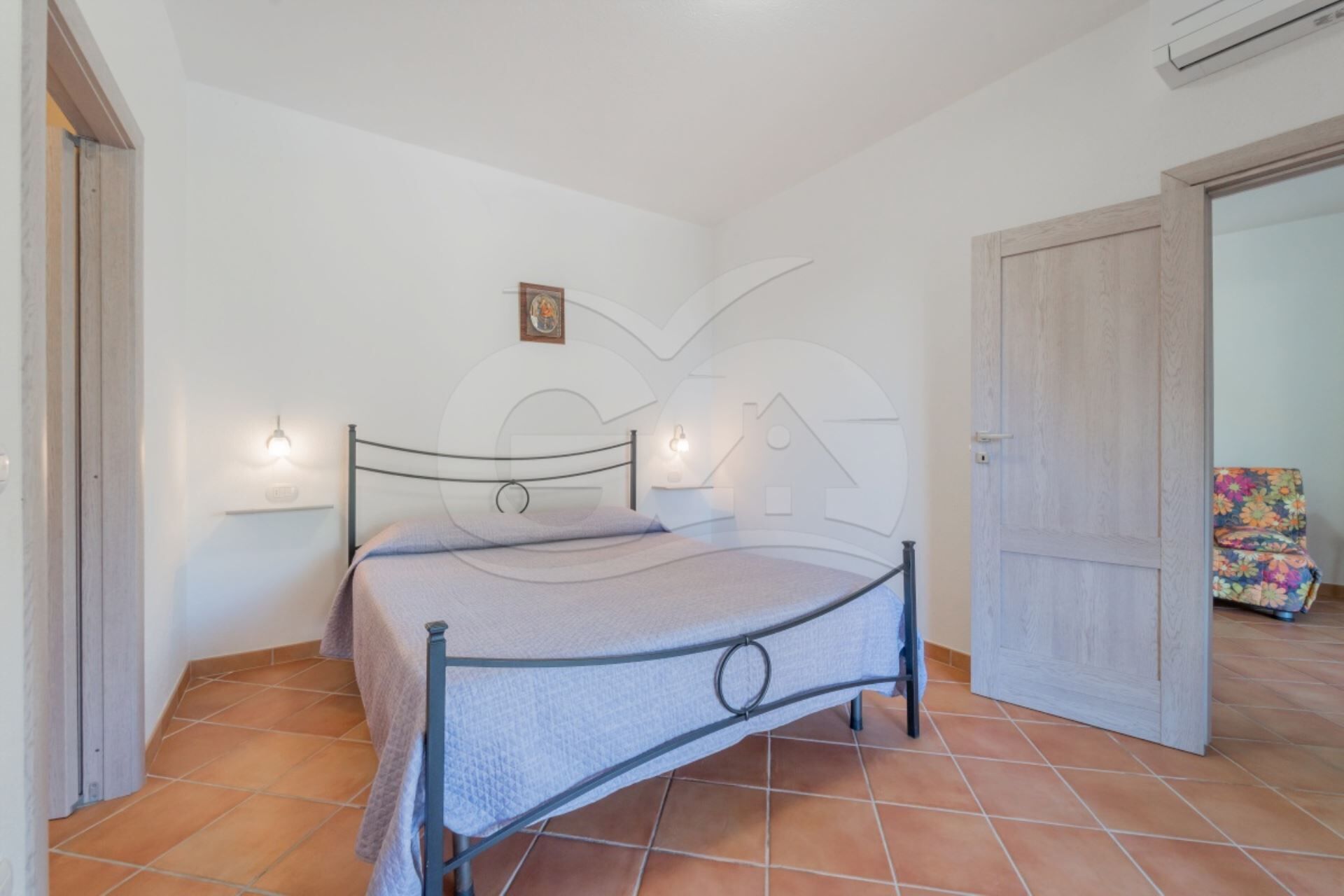 A bedroom with a bed, two wall-mounted lights, a door, and a tiled floor.