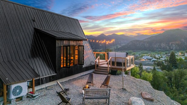 A modern building with a sloped roof, outdoor seating area with chairs and a fire pit, and a view of a valley with lights in the distance. - Cozy Aframe Cabin Zen Near Zion (Glendale)