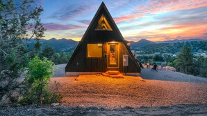 A triangular wooden cabin with a glass door, surrounded by gravel, trees, and a mountainous backdrop. - Cozy Aframe Cabin Zen Near Zion (Glendale)