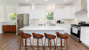 A modern kitchen with white cabinets, stainless steel appliances, and a central island with bar stools. - Sojourn on Lea View | Willoughby Spit | 5 BR *NEW* (Norfolk)