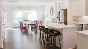 A modern kitchen with white cabinetry, a central island with stools, and a dining area with red chairs. - Sojourn on Lea View | Willoughby Spit | 5 BR *NEW* (Norfolk)