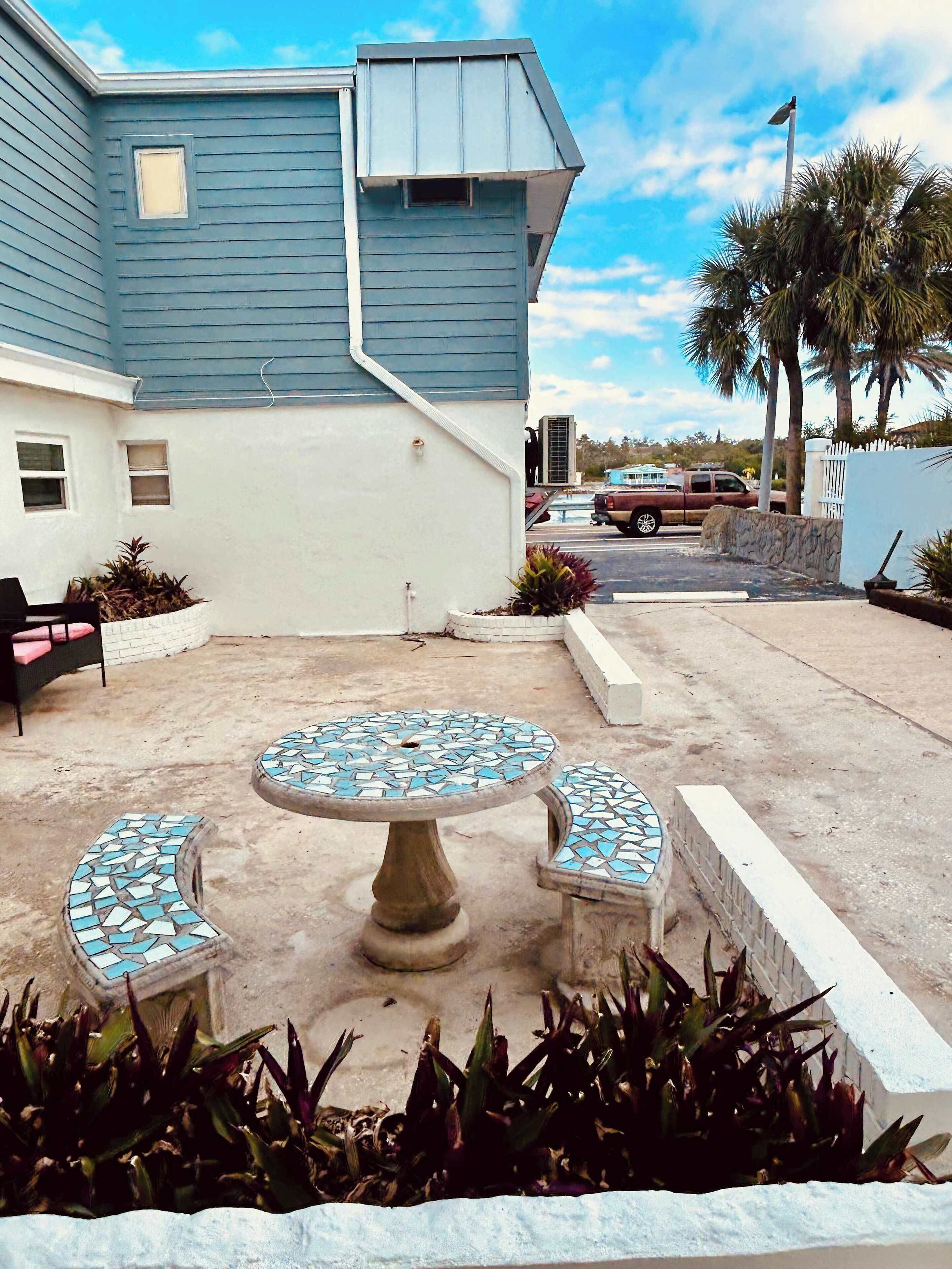A patio with a mosaic table and benches, surrounded by plants and a house with blue siding.