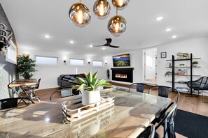 Una sala de estar moderna con una mesa de comedor de madera, sillas negras, una mesa con cubierta de vidrio, un ventilador de techo y una chimenea.