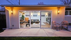 Il s’agit d’une maison moderne dotée d’une grande porte-fenêtre coulissante qui donne sur le salon. À l’intérieur, on trouve un canapé, une table basse et un téléviseur. La maison a un toit plat et une terrasse couverte avec des chaises.