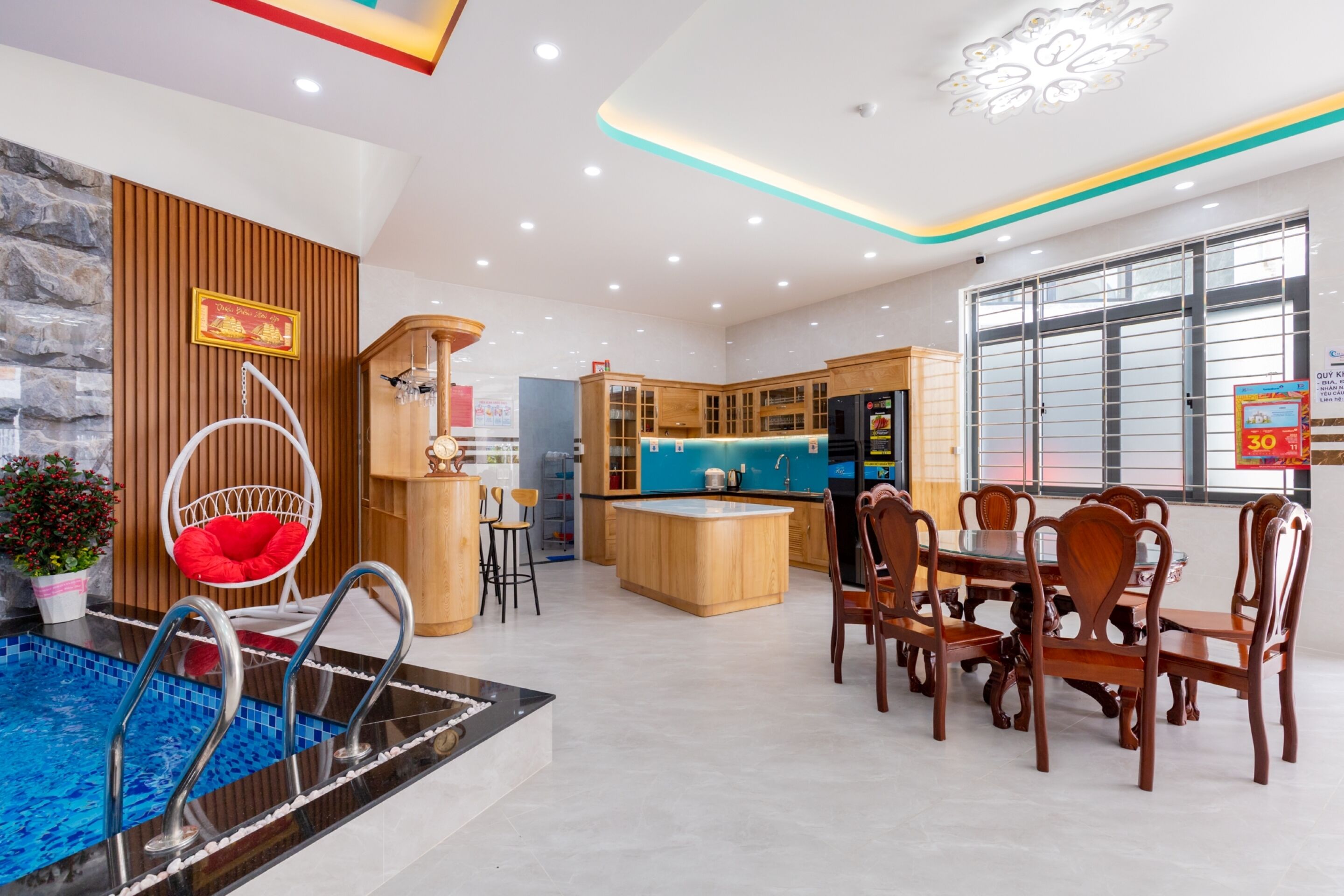 A modern kitchen with wooden cabinets and a central island, a dining area with wooden chairs, and a pool area with a red chair.