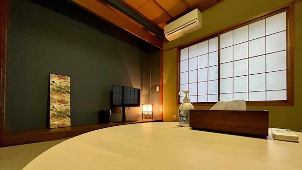 Una habitación japonesa tradicional con suelo de tatami, una mesa baja de tatami, un televisor, un dispensador de gel antibacterial y una ventana con celosías shoji.