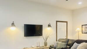 A modern living room with a flat-screen TV, two wall-mounted lamps, a ceiling fan, a leather sofa, a coffee table, and a dining area with a chair and table. - A clean, comfortable space just off the interstate with easy parking. (Worden)