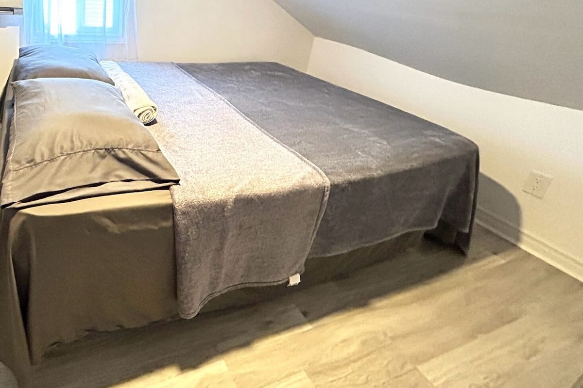 A small, single-bed room with a grey bedspread and pillows, wooden flooring, and a window with curtains.