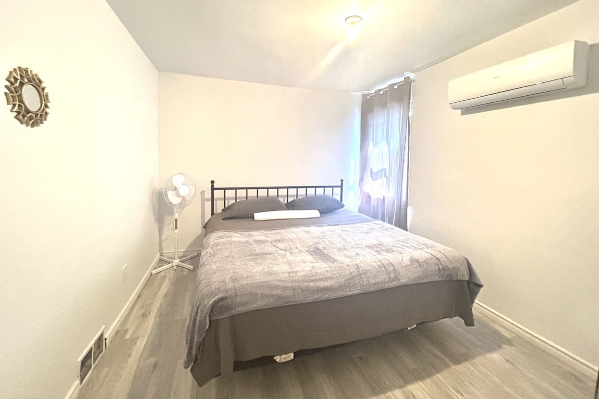 A bedroom with a bed, a bedside table, a wall-mounted air conditioner, and a window with curtains.