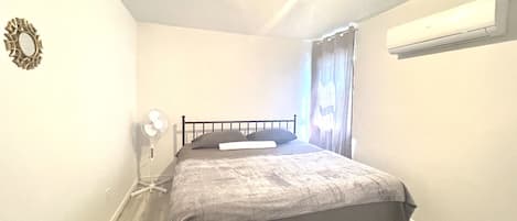 A bedroom with a bed, a bedside table, a wall-mounted air conditioner, and a window with curtains.