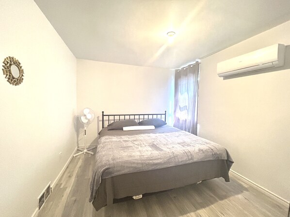 A bedroom with a bed, a bedside table, a wall-mounted air conditioner, and a window with curtains. - Aubin Paradise (Windsor)