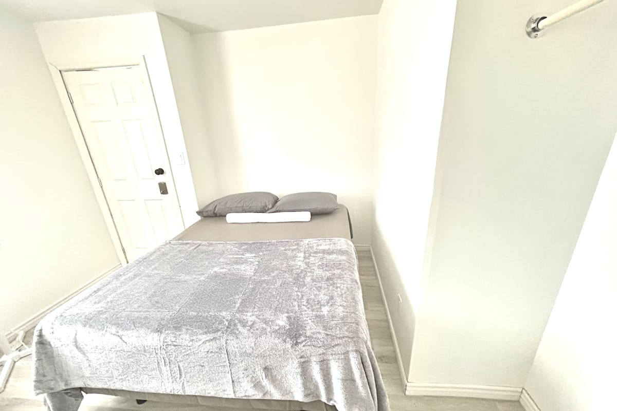 A small, single-bed room with a grey bedspread, two pillows, and a white headboard. A door is visible on the left, and a curtain rod is on the right.