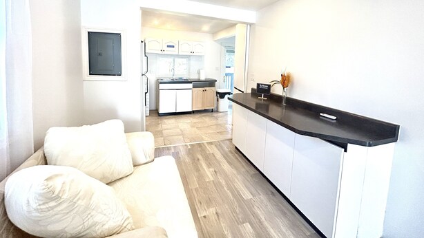 A modern kitchen with white cabinets and a black countertop, a white sofa with cushions, and a view into the dining area. - Aubin Paradise (Windsor)