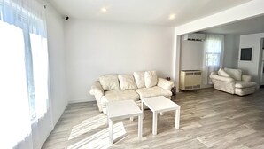 A spacious living room with a light-colored sofa, two white side tables, and a large window with sheer curtains. - Aubin Paradise (Windsor)