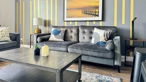 A modern living room with a grey sofa, a coffee table, and a framed painting of a sunset over a pier. - Stylish 2BR Apartment with City View (Atlanta)
