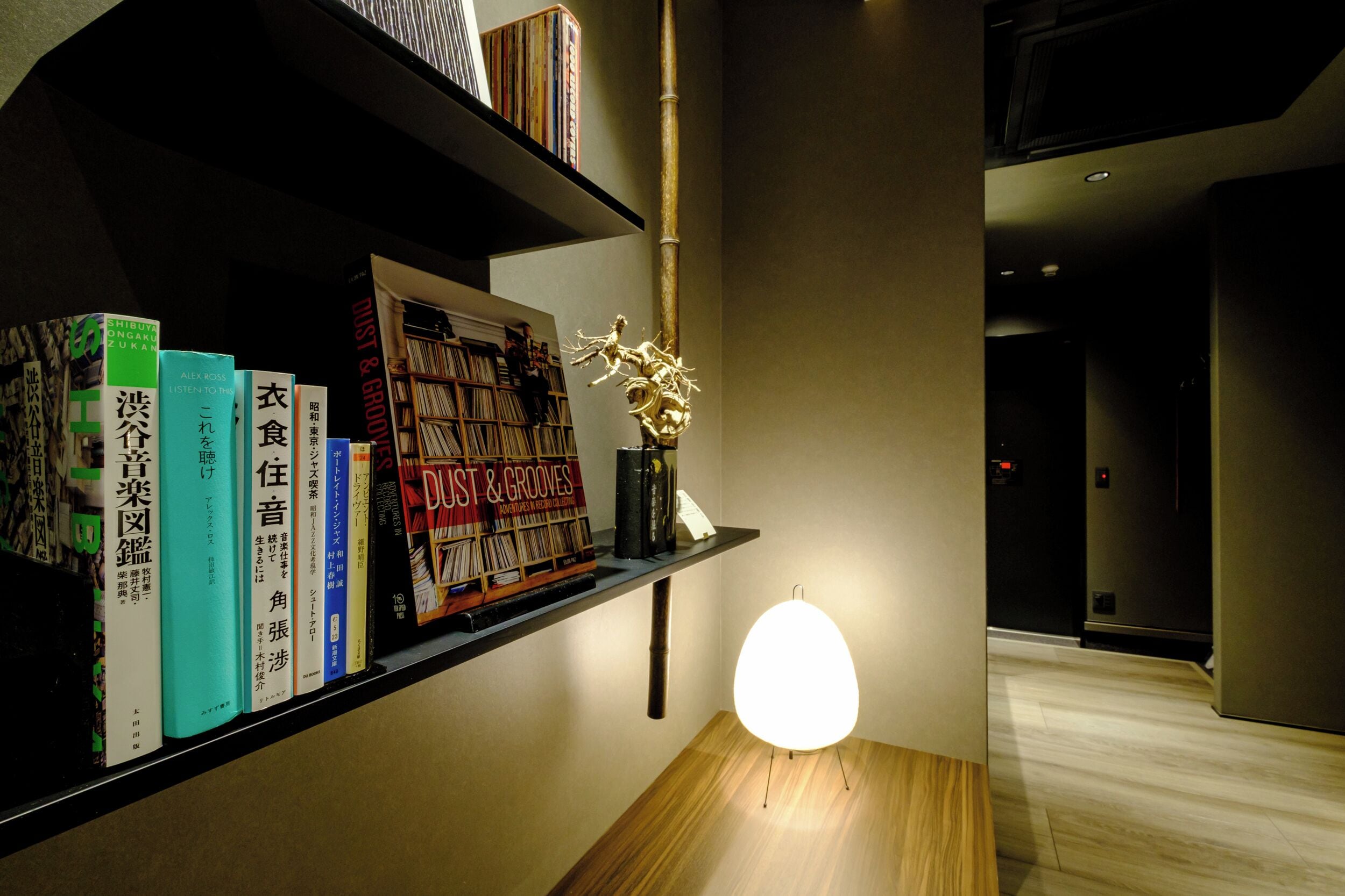 A hallway with a wooden floor, a shelf with books, a decorative vase, and a wall-mounted light fixture.
