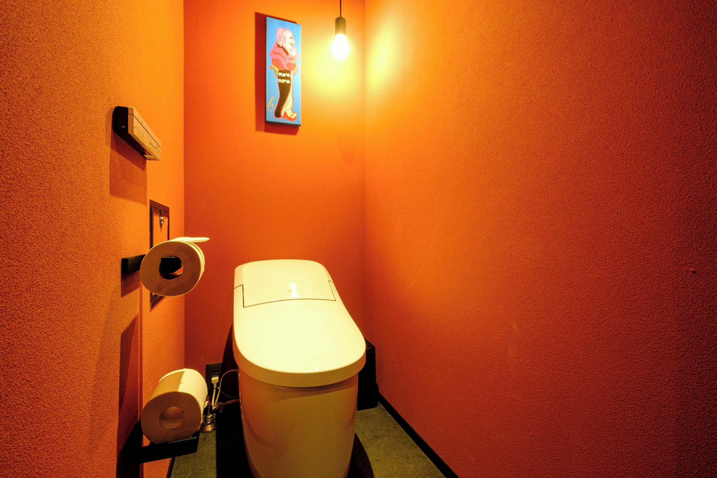 A small bathroom with an orange wall, a toilet, a roll of toilet paper, and a light fixture.