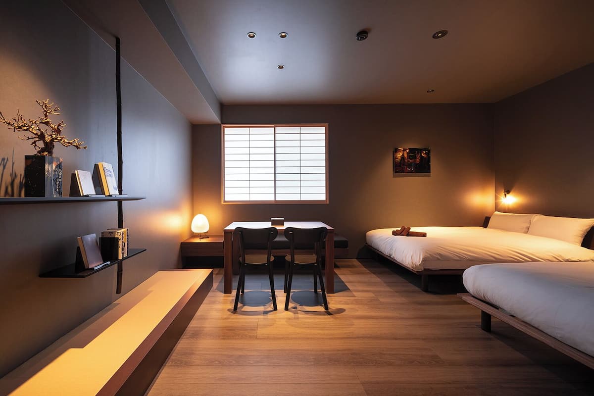 Un dormitorio moderno con escritorio, dos sillas, cama, ventana con persianas y estantería en la pared con libros y una planta.