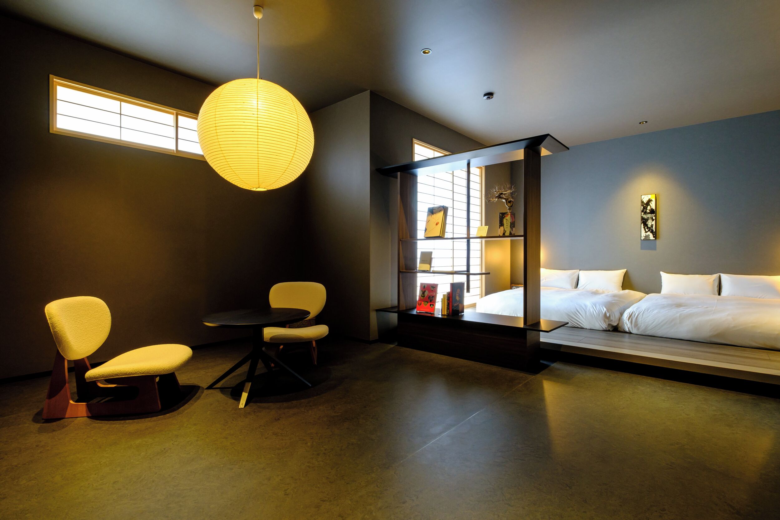 A modern bedroom with a bed, two chairs, a small table, a bookshelf, and a large window.