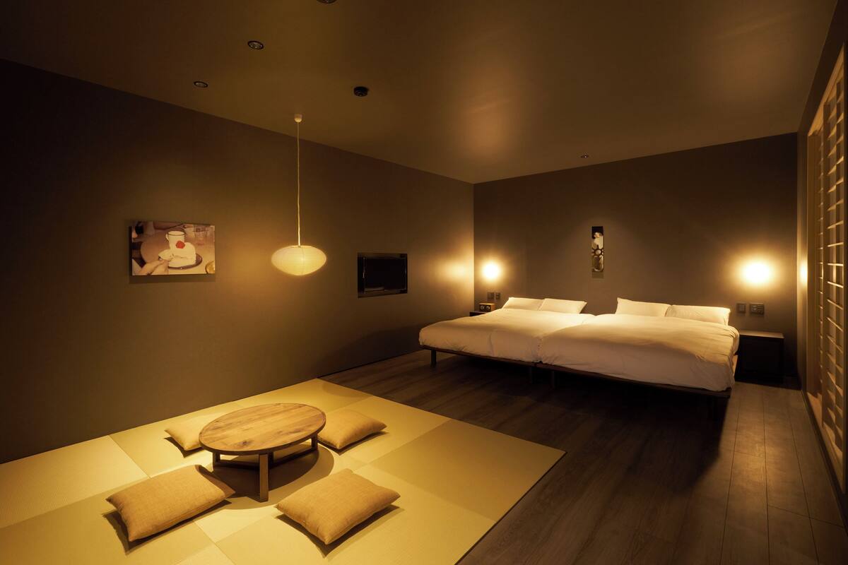 Una habitación de hotel moderna con una cama grande, una mesita con cojines y un televisor montado en la pared.