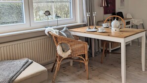 A cozy room with a large window, a dining table with chairs, a wicker chair with a fur throw, and a radiator. - Hygge Frisian House by the Sea with Private Garden and Terrace (Nordstrand)
