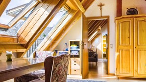 Une pièce avec un plafond en pente, des poutres en bois, une table à manger, une chaise, un buffet et une croix accrochée au mur.