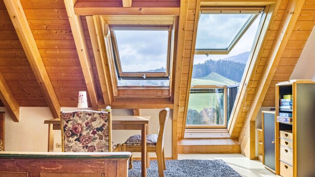 Une pièce avec un plafond en pente, des poutres en bois, une lucarne, une table, une chaise et un fauteuil à motifs floraux, ainsi qu’une bibliothèque.
