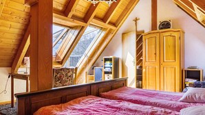 Une chambre avec un plafond en pente, des poutres en bois, un lit avec une literie rose, une armoire et un ventilateur.