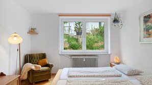 A bedroom with a bed, a chair, a floor lamp, a radiator, a window, and a hanging plant. - Apartment 'Ferienwohnung Am Seebach, 80 Qm' with Private Terrace and Wi-Fi (Titisee-Neustadt)