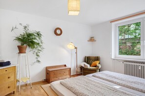 A bedroom with a bed, a chair, a dresser, a plant, a lamp, and a window.