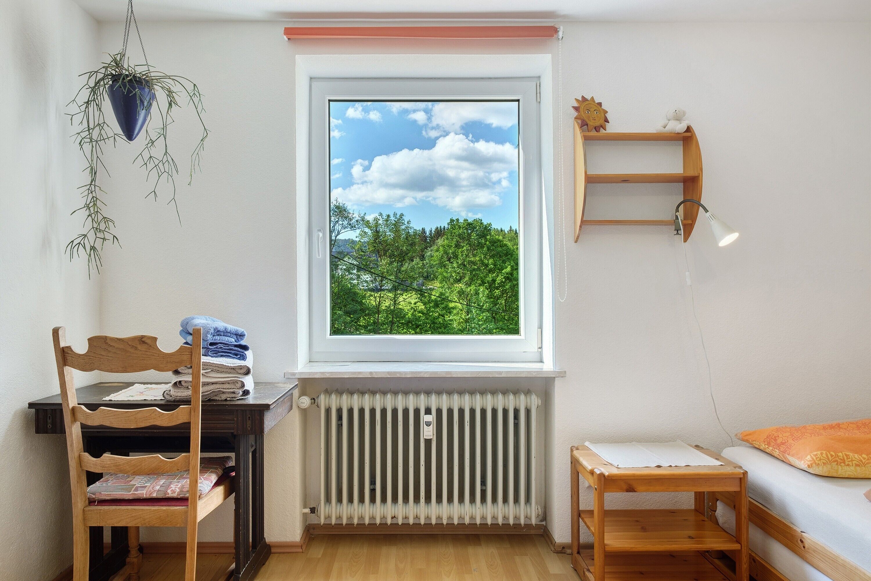 Quarto com janela, radiador, cadeira de madeira, mesa pequena e planta pendente.