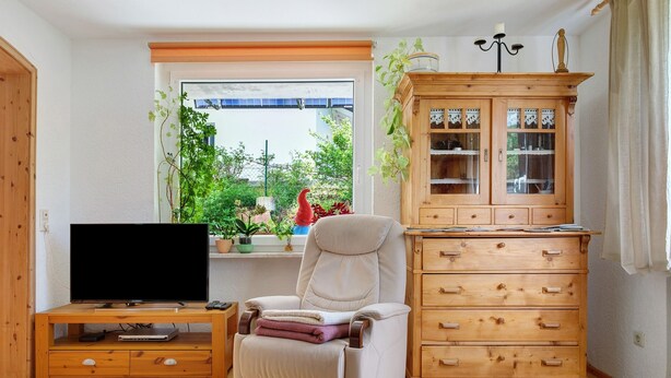A living room with a TV on a wooden stand, a recliner chair, a wooden cabinet, and a window with a view of greenery. - Apartment 'Ferienwohnung Am Seebach, 80 Qm' with Private Terrace and Wi-Fi (Titisee-Neustadt)
