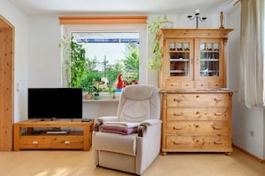 Un salon avec un téléviseur posé sur un meuble en bois, un fauteuil inclinable, une armoire en bois et une fenêtre donnant sur un paysage verdoyant.