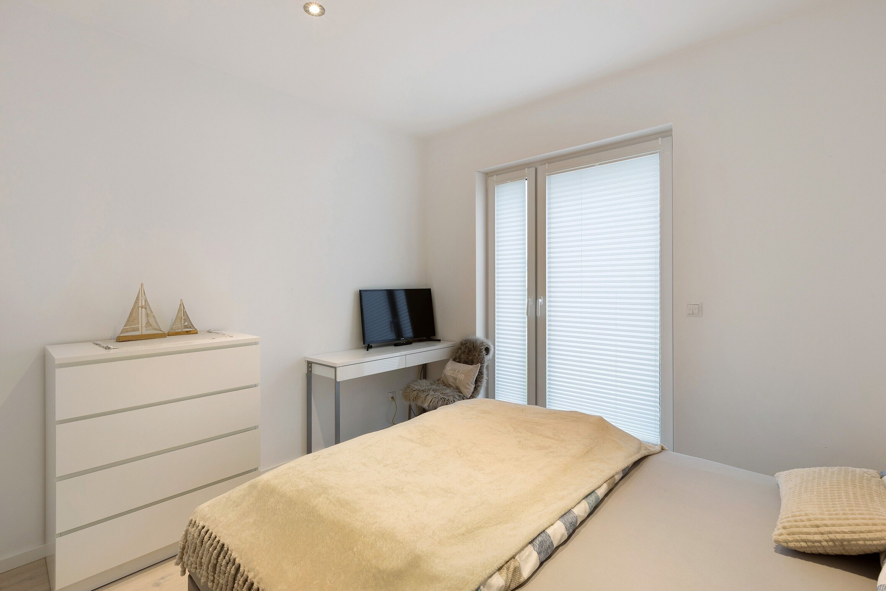 Un dormitorio con cama, cómoda, escritorio con televisor y ventana con persianas.