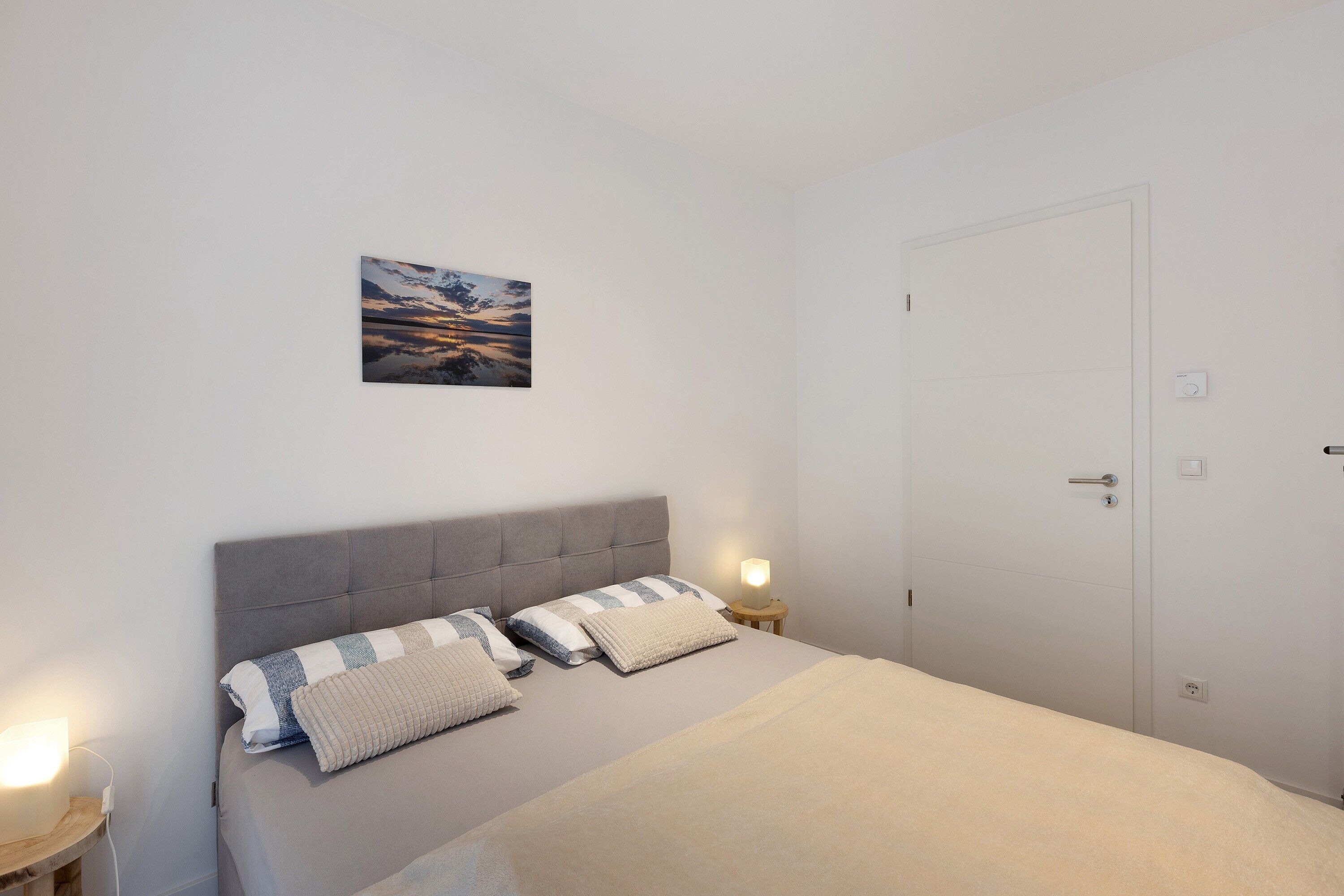 Un dormitorio con cama, mesitas de noche, una puerta y un cuadro colgado en la pared.