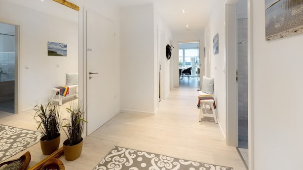Un couloir avec un plafond orné de poutres en bois, des murs blancs et une vue sur un coin repas comprenant une table et des chaises blanches.