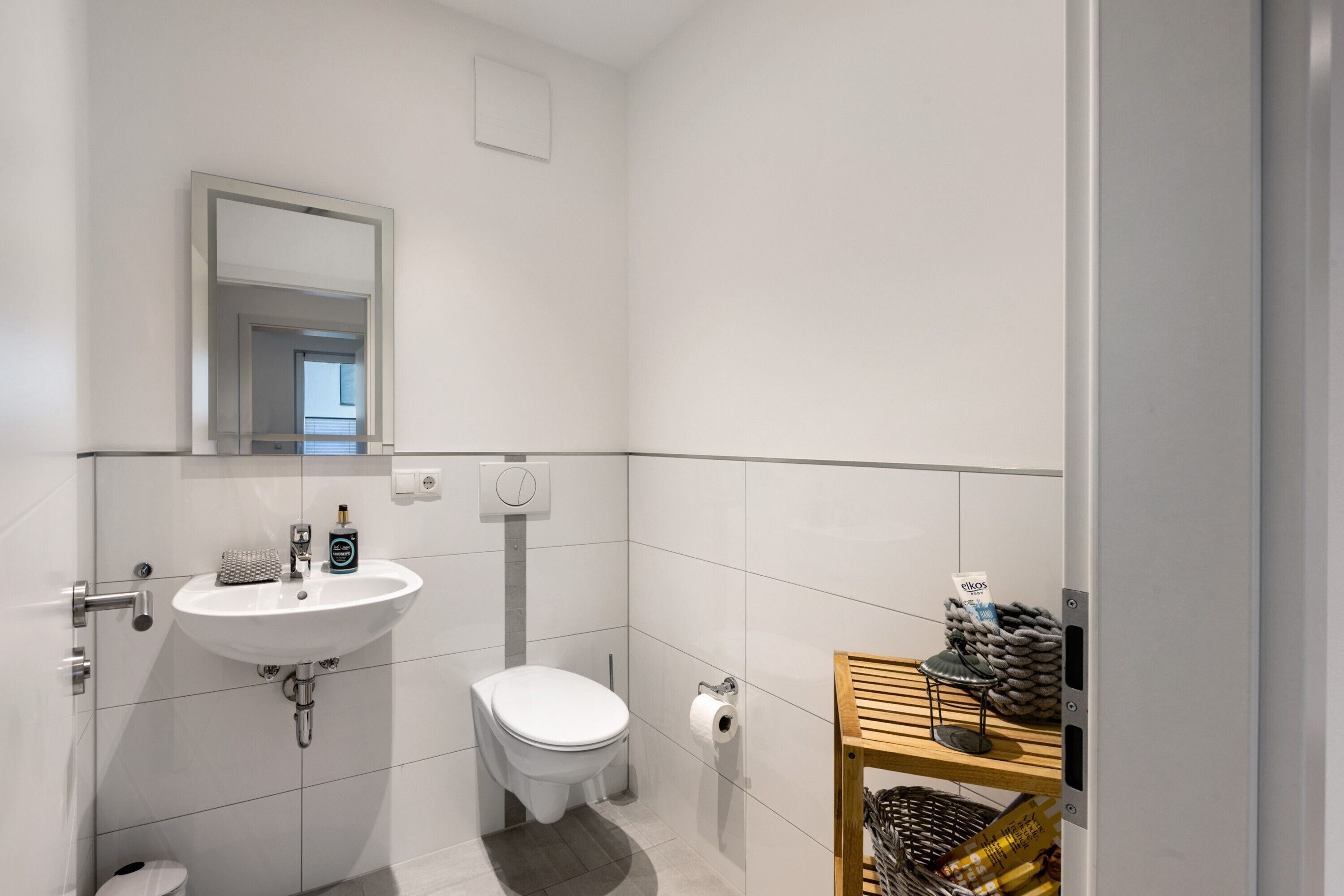 A modern bathroom with a white sink, toilet, and mirror. There is a wooden shelf with baskets and a small table.