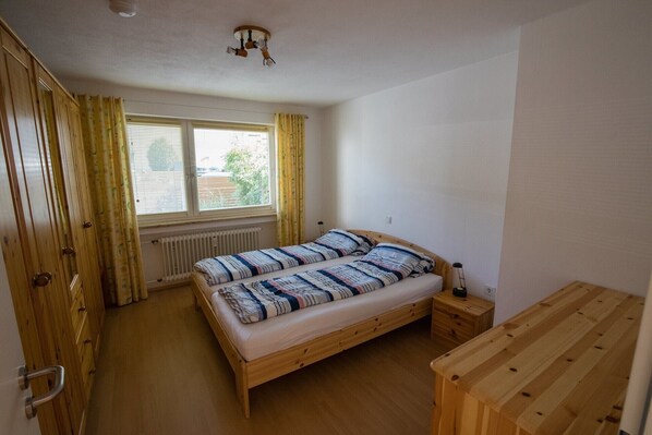 Un dormitorio con una cama de madera, una mesita de noche pequeña, una ventana con cortinas y un radiador.