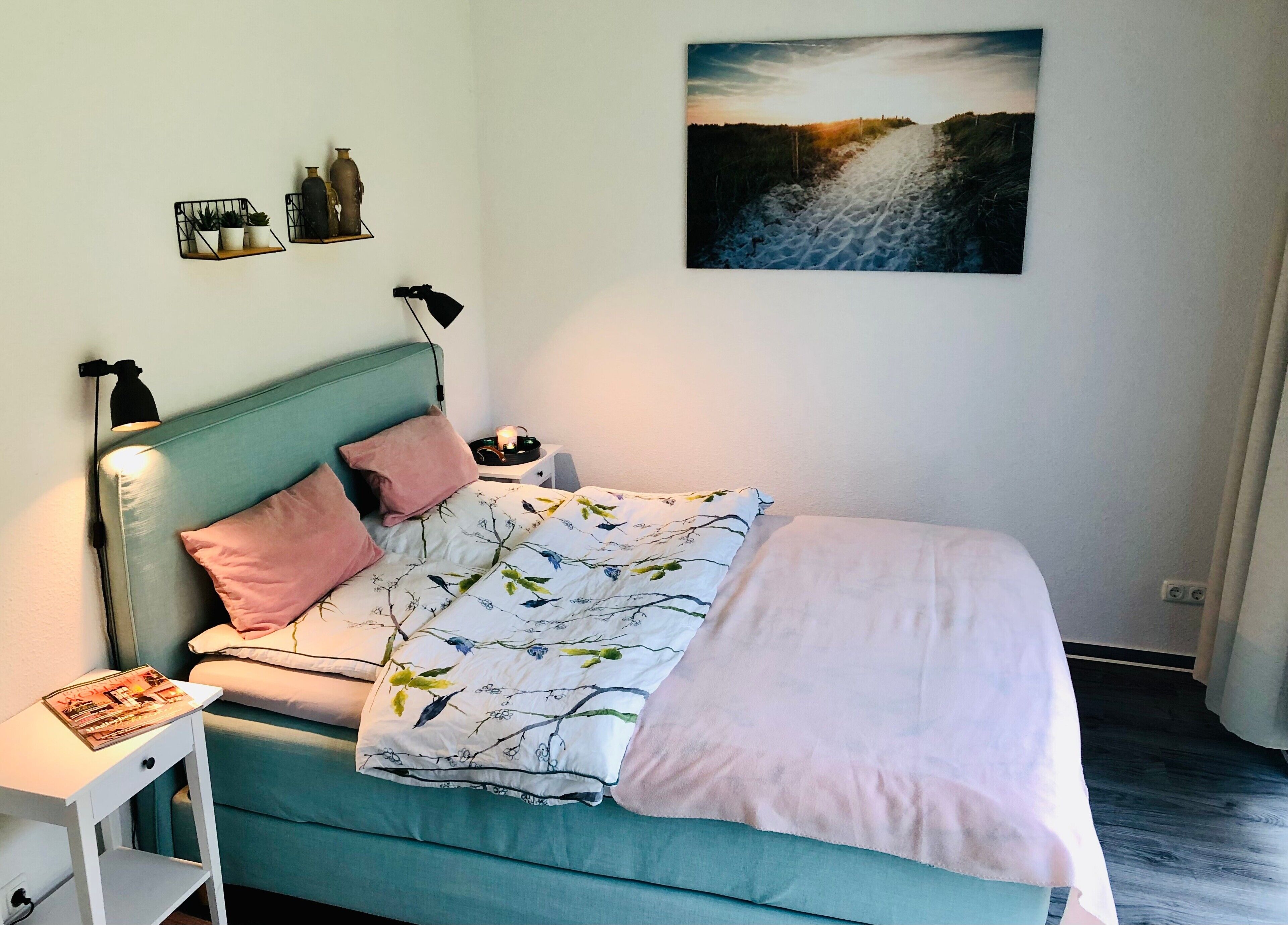 Un dormitorio con cama, mesita de noche, lámpara, estante en la pared con plantas y un cuadro.