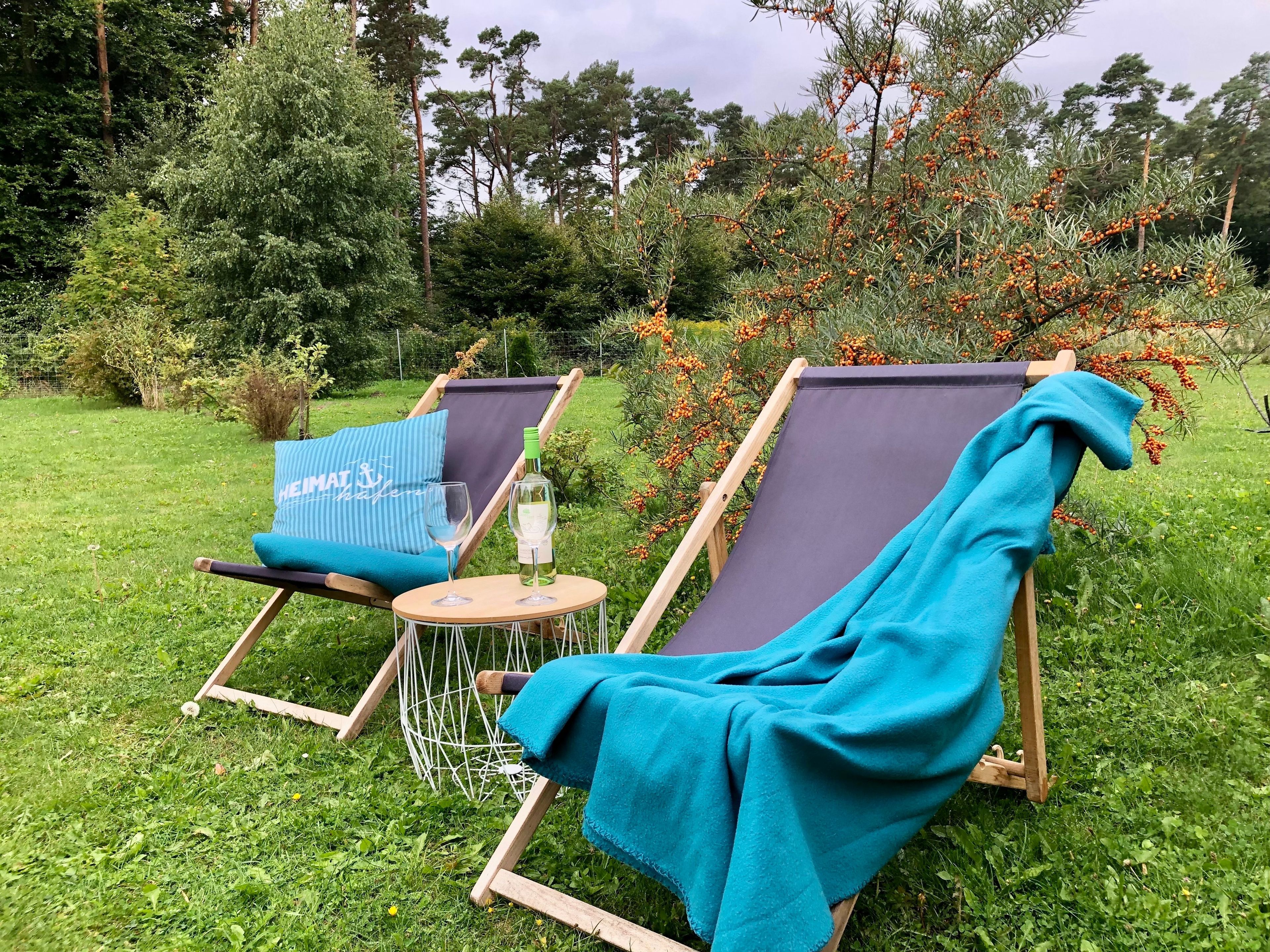 Due poltrone con cuscini, un tavolino rotondo e una bottiglia di vino in un'area verde all'aperto con alberi e arbusti.