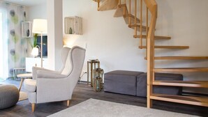 Un salon moderne avec un escalier en bois, un canapé gris, un pouf rond et une lampe.