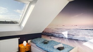 A bedroom with a sloped ceiling, a window, a bed with a blue patterned bedspread, a bedside table with a lamp, and a radiator.